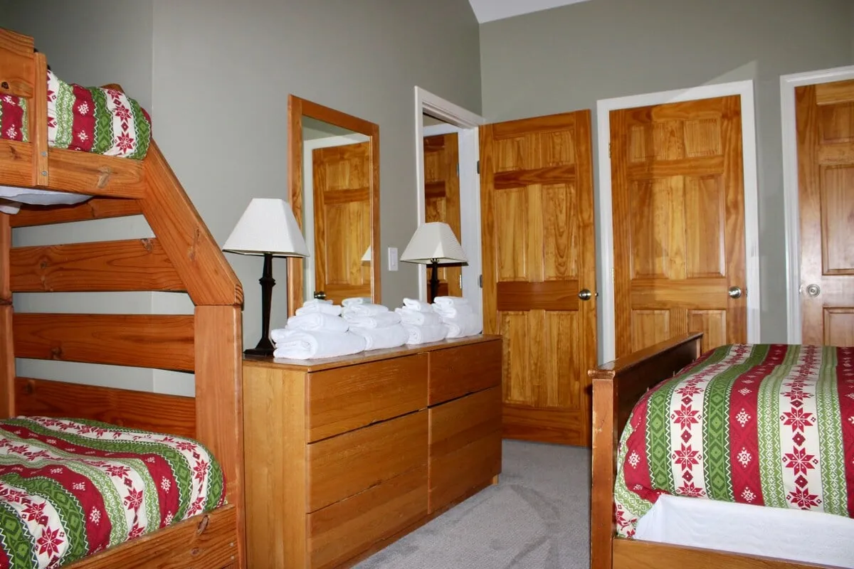 A cozy bedroom with a double-over-double bunk bed, a sturdy dresser, soft lamps, and playful, decorative bedding. The layout is spacious enough for group or family stays, and the mood is warm and inviting, suggesting shared memories in a welcoming environment.