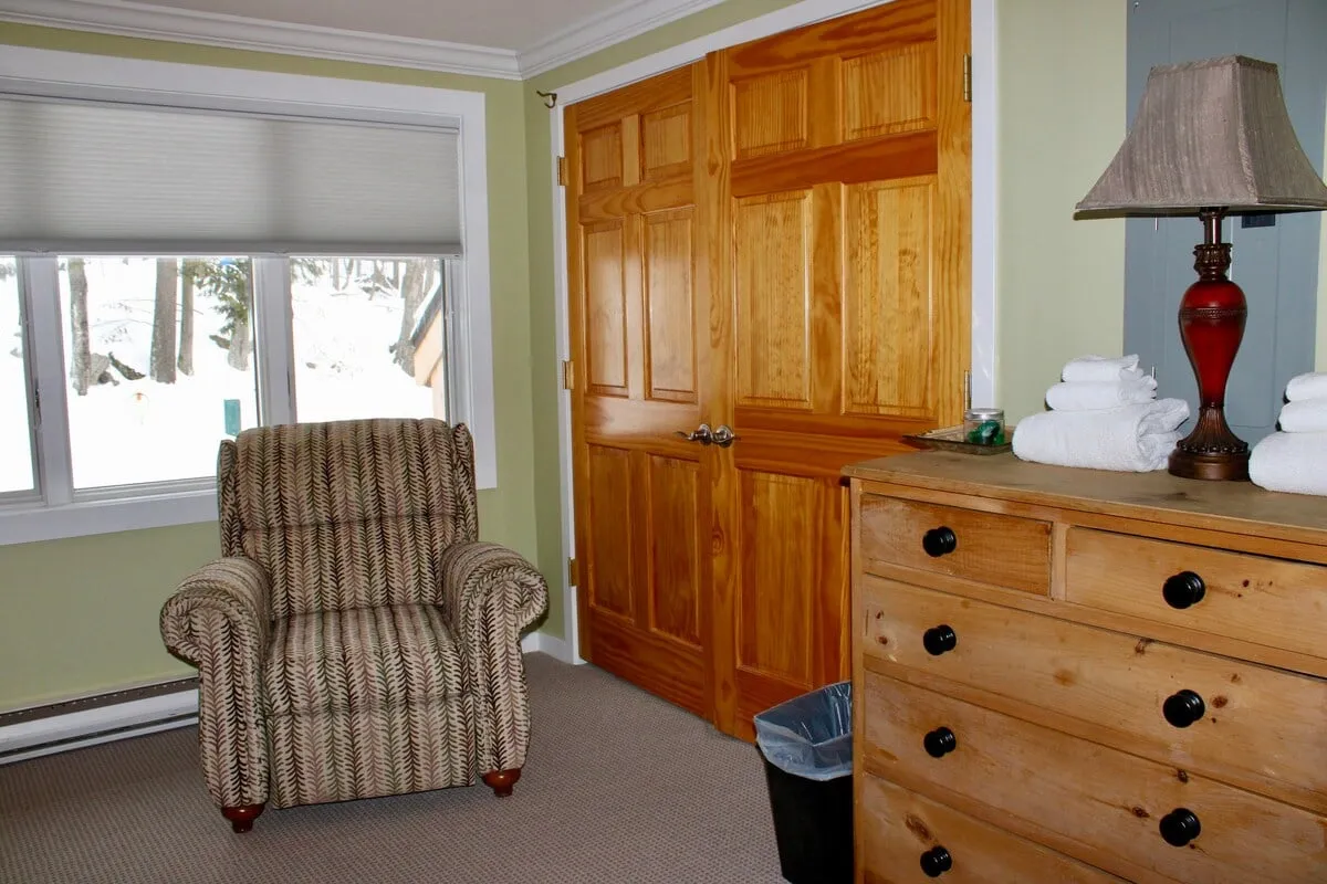 A cozy interior nook with a striped armchair, wooden dresser, lamp, and neatly stacked towels. The mood is relaxed, perfect for unwinding with a book or a hot drink after a busy day.