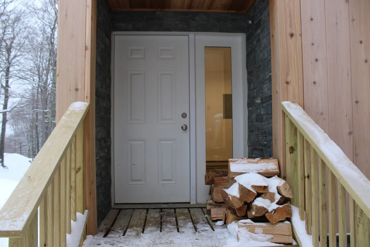 The welcoming entryway of the home with a stack of firewood, wooden railing, and the front door framed by winter scenery. The scene suggests both arrivals after a day on the slopes and the warmth that awaits inside.