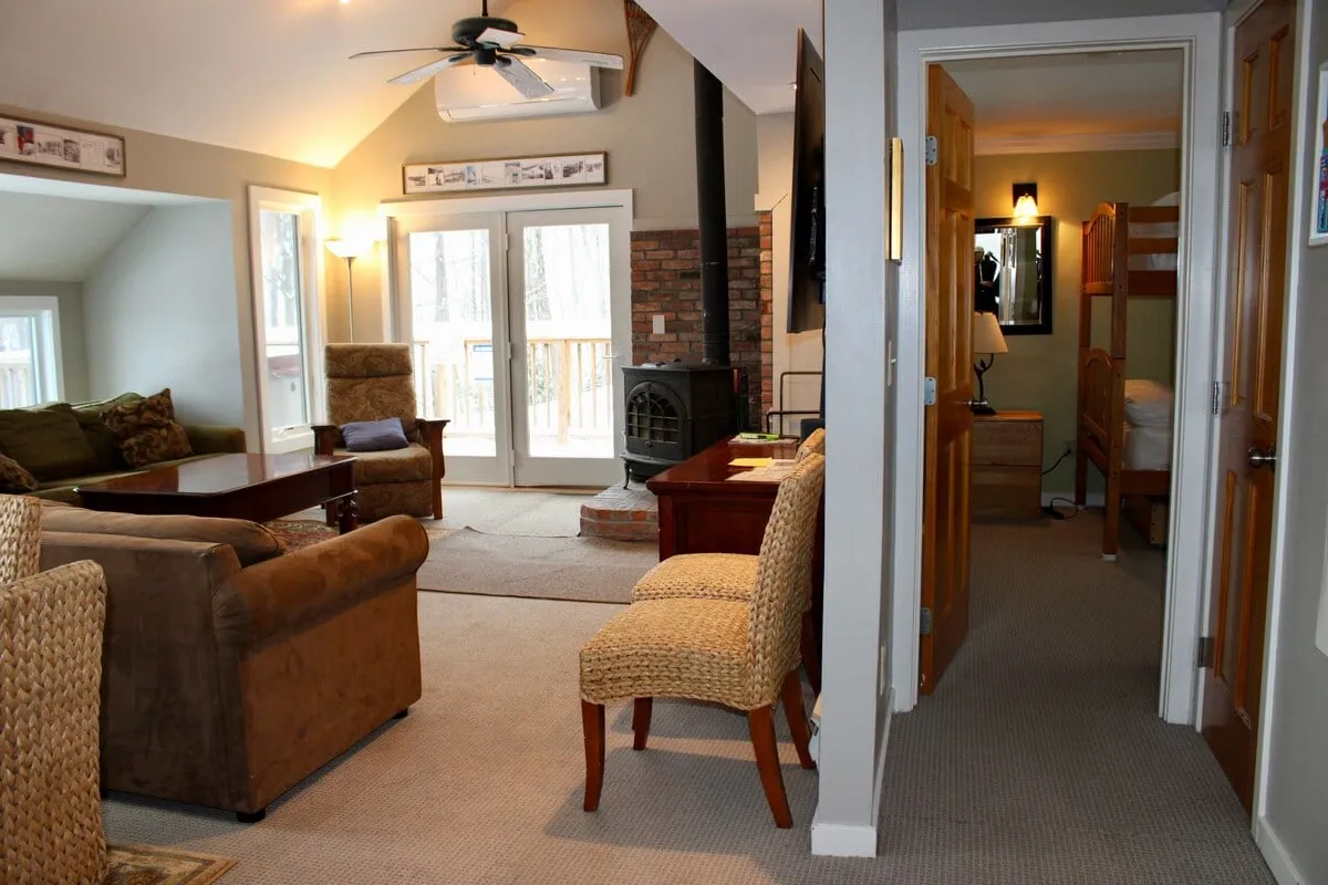 This image depicts a spacious, open-plan living room with a comfortable sofa, wooden coffee table, and a cozy fireplace. The dining area is visible in the background, and bunk beds add a family-friendly touch. The scene is lively and warm, with people relaxing, socializing, and possibly playing games.