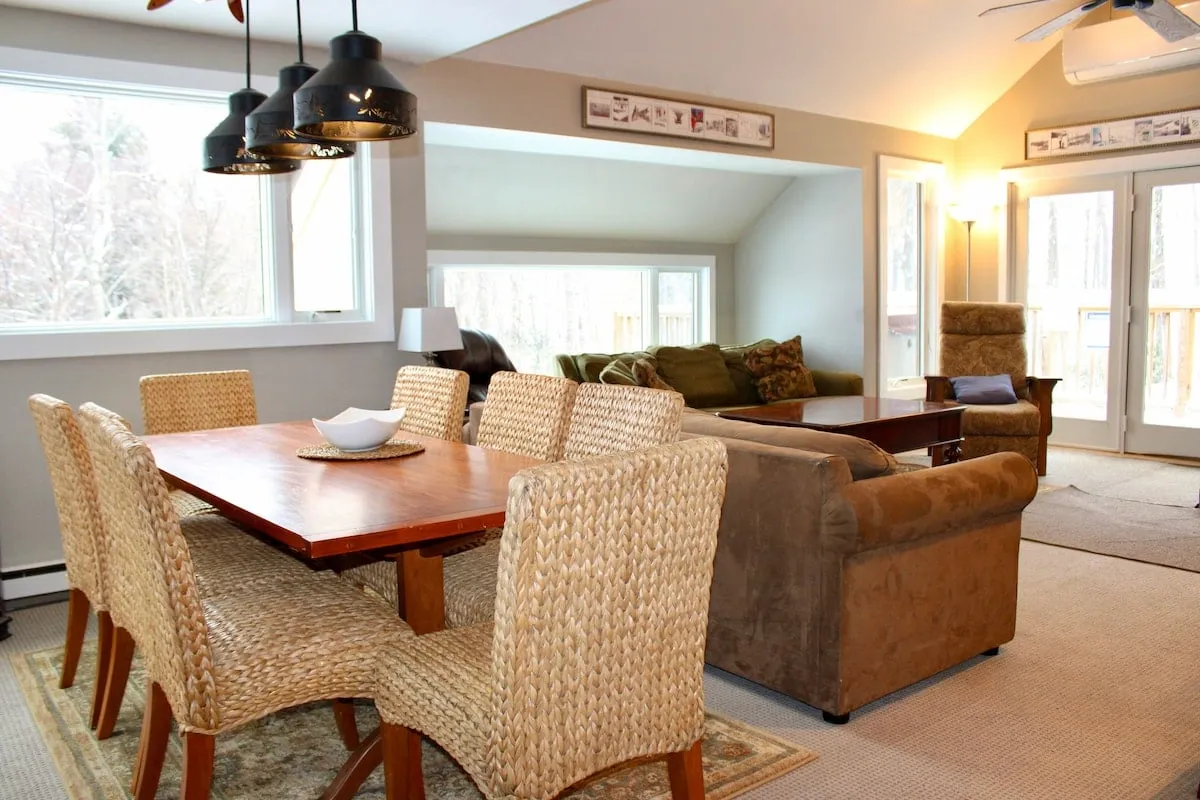 The image showcases a warm, inviting living and dining area with a comfortable sofa and a spacious dining table, all bathed in natural light from large windows that overlook the picturesque Vermont landscape. The setup is perfect for cozy family meals, relaxing with a book after a day on the slopes, or gathering with friends for games and social time.