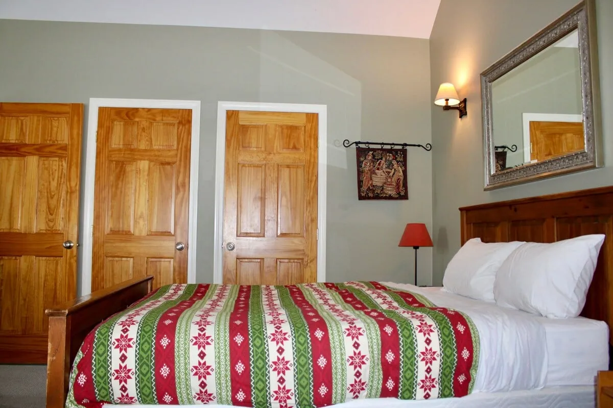 This image features a cozy bedroom with a patterned duvet bed, warm wooden doors, a decorative wall hanging, a mirror, and a softly glowing table lamp. The scene evokes a sense of relaxation and comfort, suggesting a peaceful retreat ideal for resting or sharing quiet moments after a busy day.