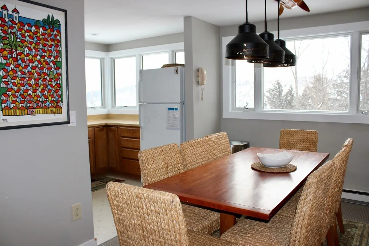 This image depicts a warm, sunlit dining area with a wooden table and woven chairs, set before large windows that offer glimpses of the outdoors. The artwork on the wall and inviting setting suggest a communal space ideal for recharging between adventures.