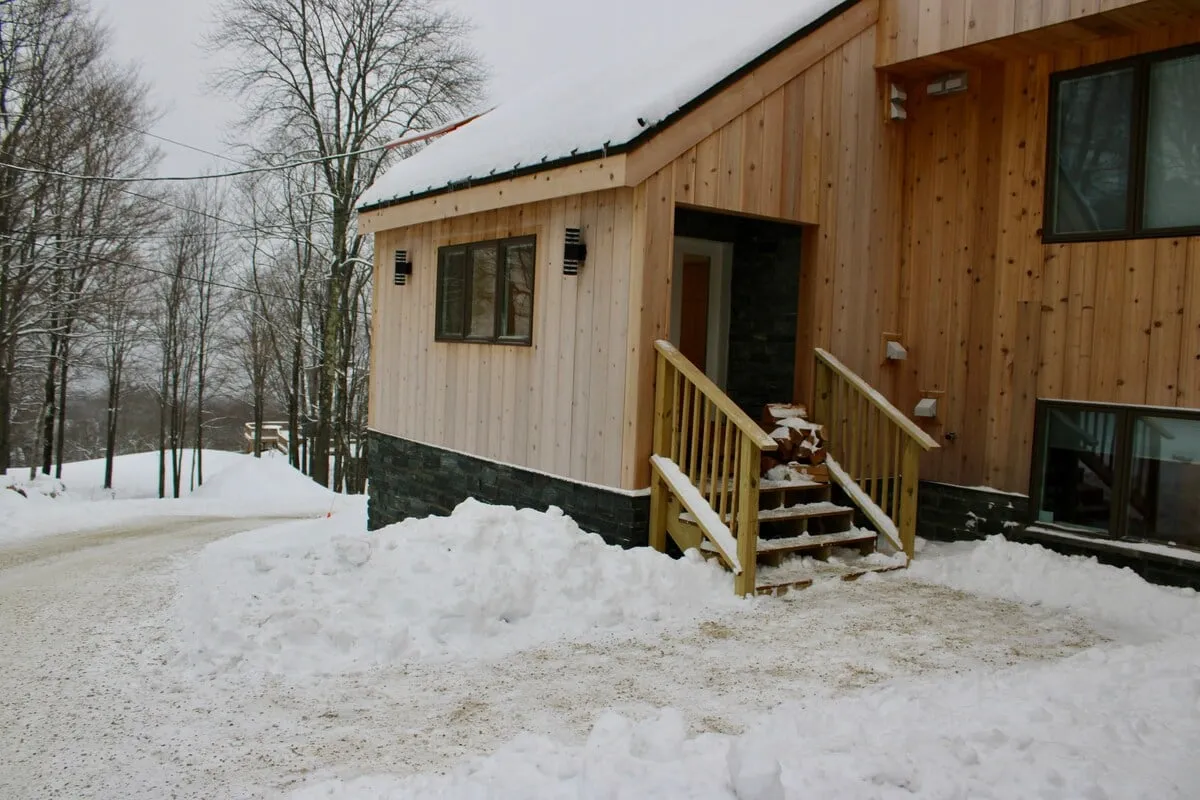 A tranquil winter exterior scene featuring the facade of the house, wooden stairs leading up to the entrance, and a snow-covered driveway set against a serene backdrop. The inviting atmosphere is enhanced by the suggestion of cozy gatherings and outdoor winter fun right outside the door.