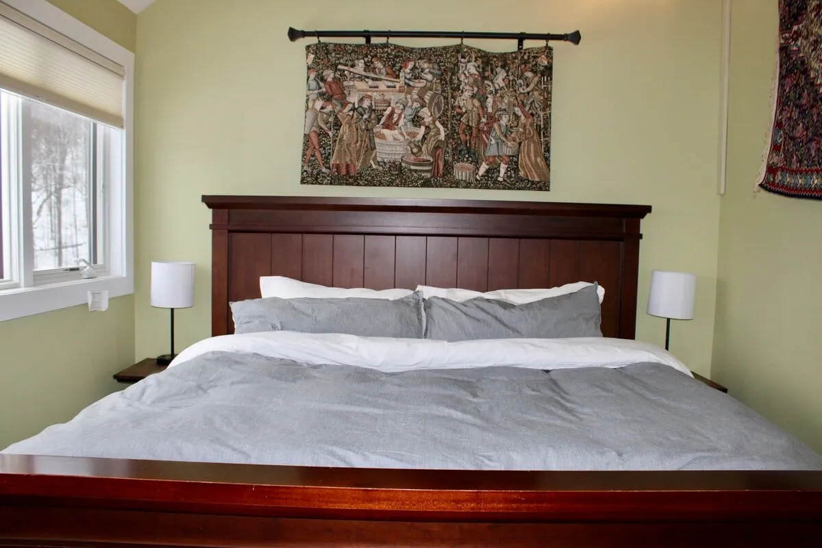 A serene bedroom scene featuring a wooden bed frame with richly textured linens, a decorative tapestry above the headboard, and soft natural light streaming in. The space feels peaceful and inviting, perfect for restful sleep or quiet relaxation.