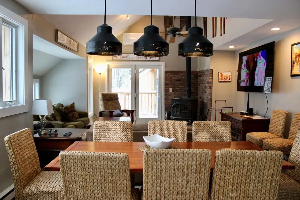 A cozy interior scene showcasing a dining table set for meals, modern light fixtures overhead, a TV, and a fireplace. The space is arranged for comfort and shared experiences, ideal for family meals or lively game nights.