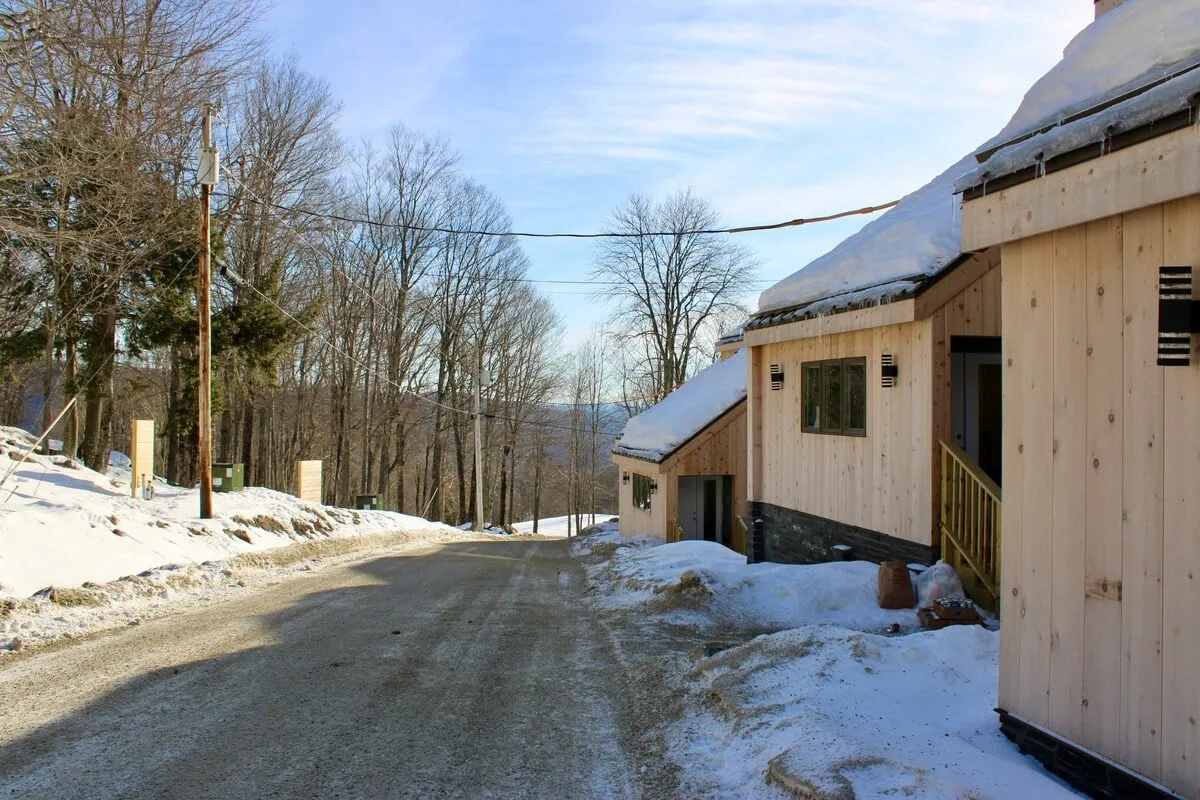 A peaceful neighborhood street lined with snow-covered houses and bare trees under a clear blue sky. The setting evokes the quiet beauty of winter and the anticipation of outdoor activities like skiing, snowshoeing, or simply enjoying nature.