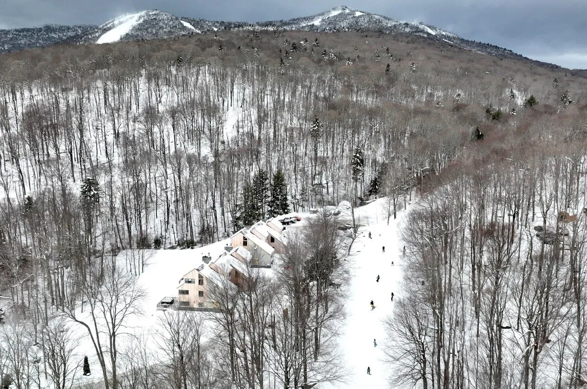 A sweeping exterior view of the property set against a dramatic, snow-covered mountain backdrop. The landscape is serene, with wooded areas and snowy expanses suggesting both privacy and immersion in the alpine environment. The property structure itself is visible, reinforcing the mountain escape atmosphere and the proximity to outdoor adventure.
